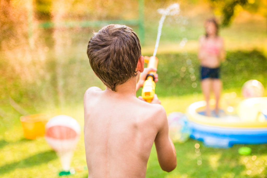 Boy splashing girl with water gun in garden swimming pool, sunny summer day, back yard,